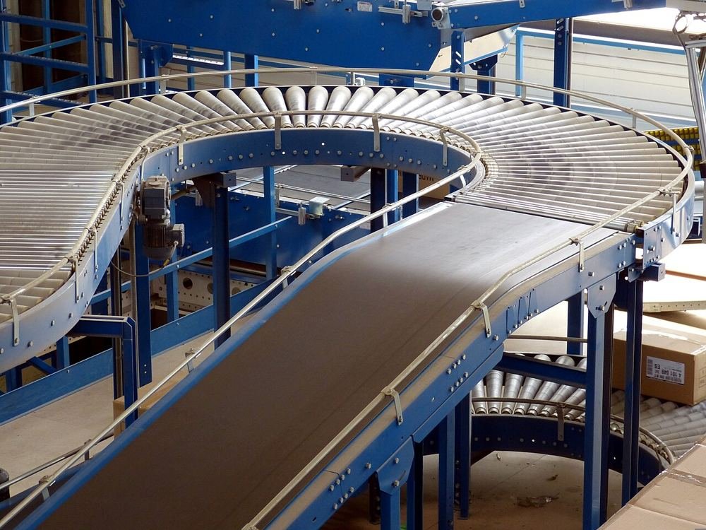 Un convoyeur de centre logistique : une bande inclinée, puis des rouleaux en courbe ; les cartons attendent à droite.